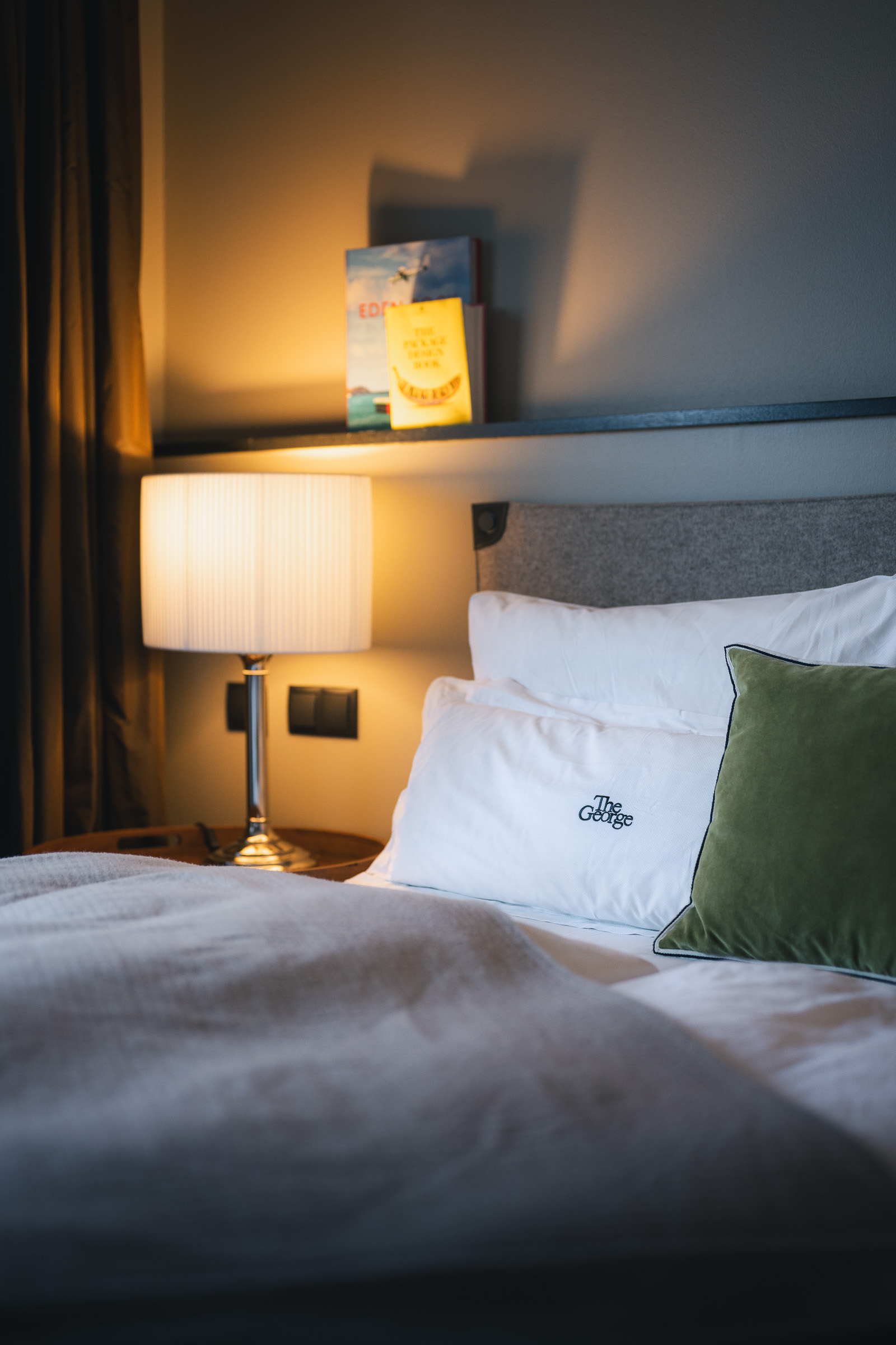 Hotel bed with white linens, a green cushion and warm bedside lighting