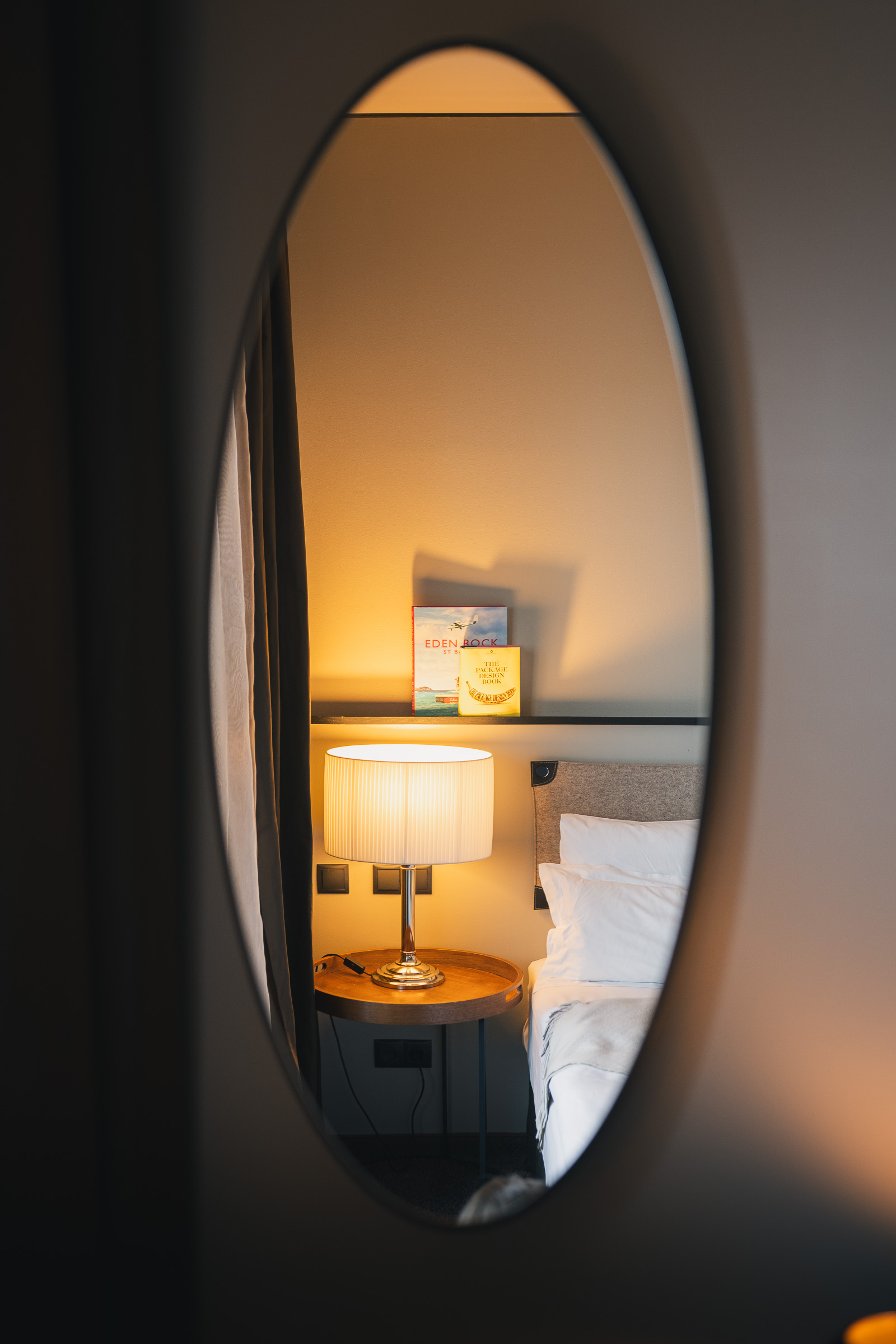 Oval mirror reflecting a warmly lit bed in a guest room