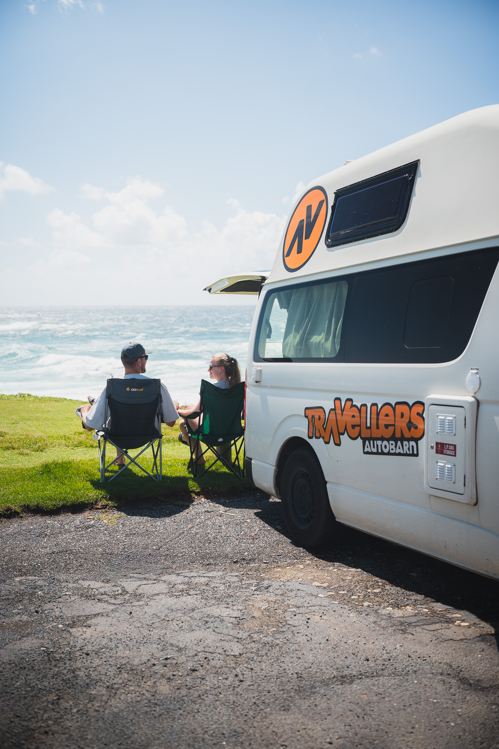 Two travelers watching the ocean from folding chairs beside their camper