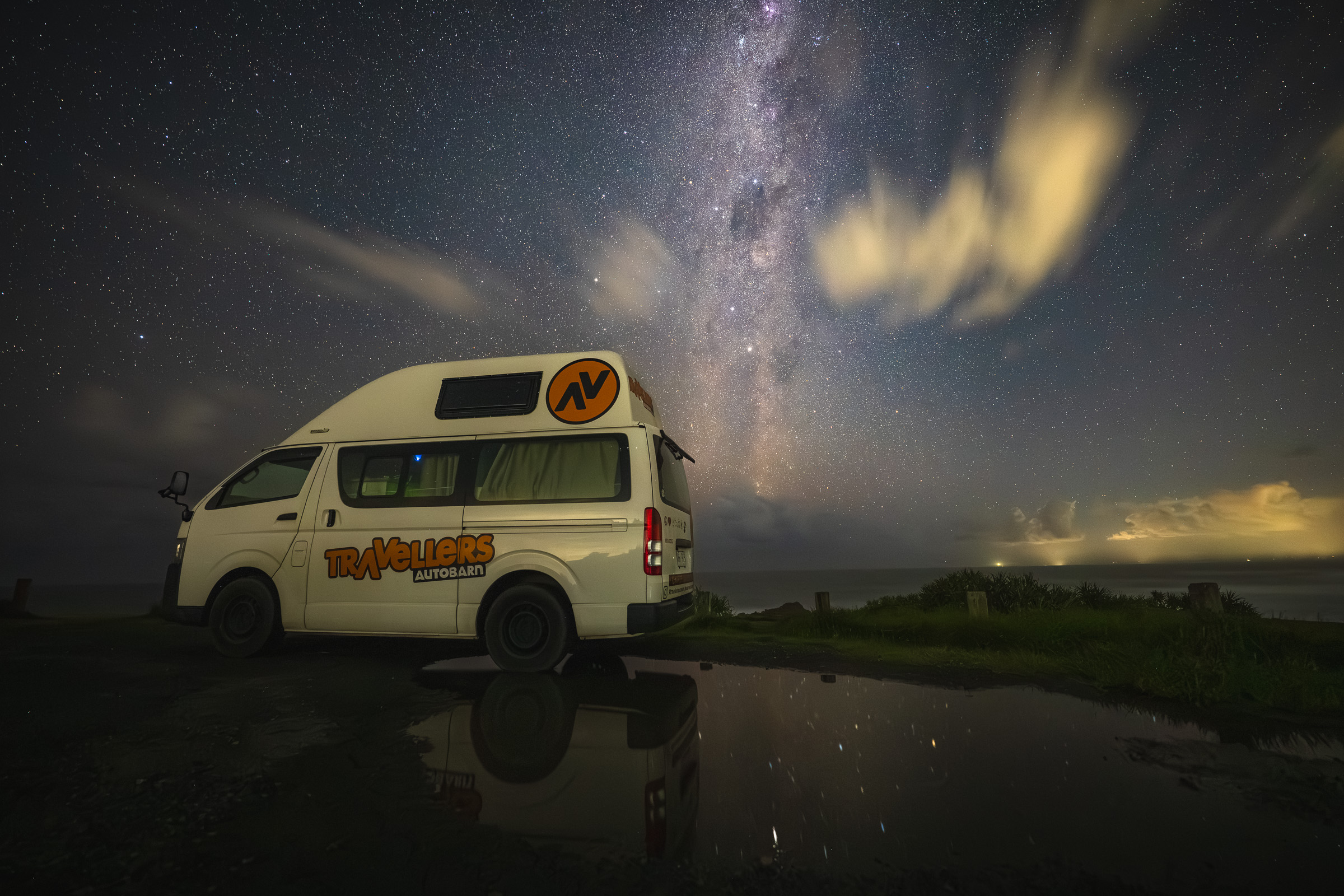 Camper van beneath the Milky Way reflected in a roadside puddle