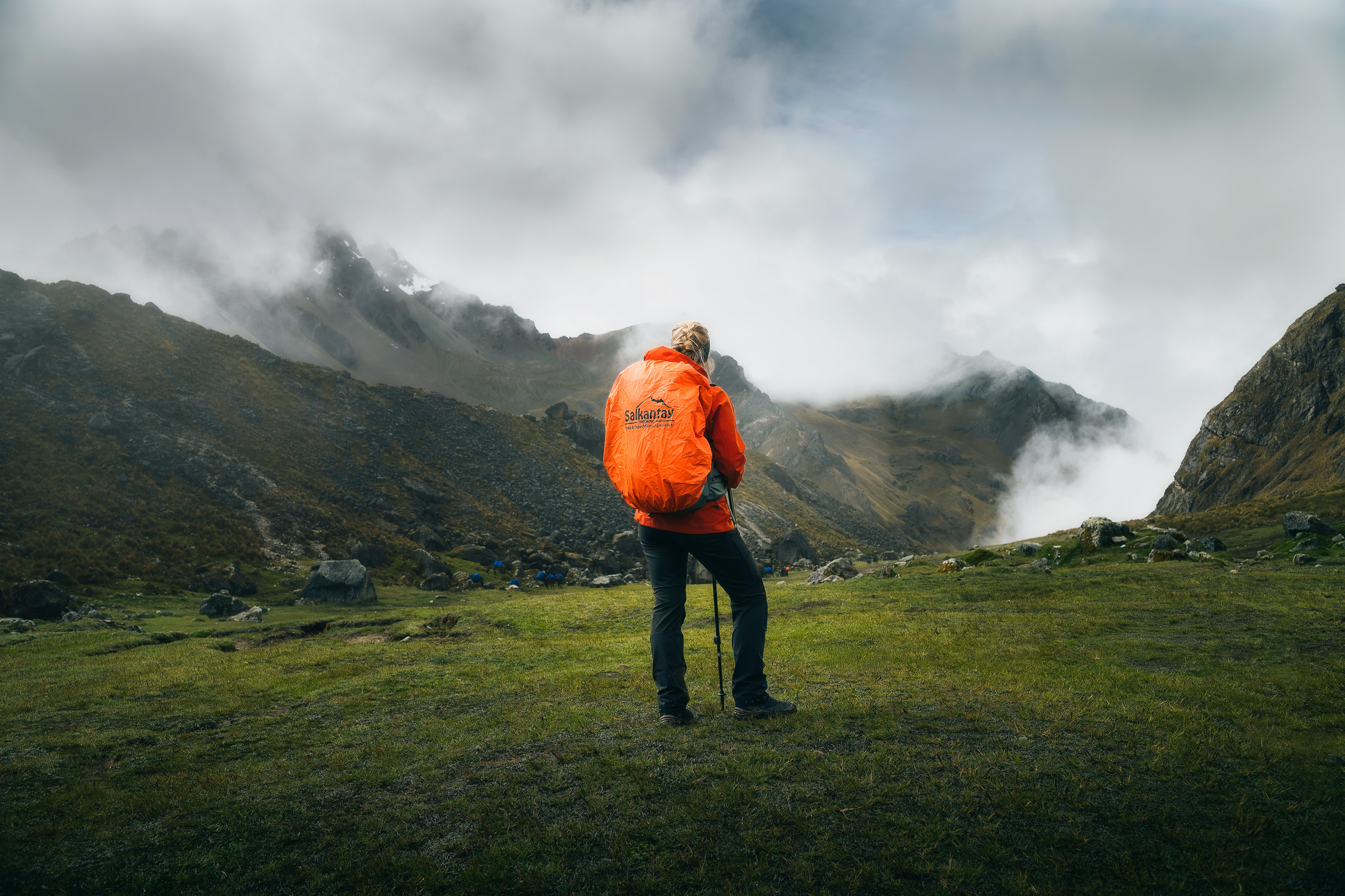 Hiker in an orange rain cover facing a misty Andean pass