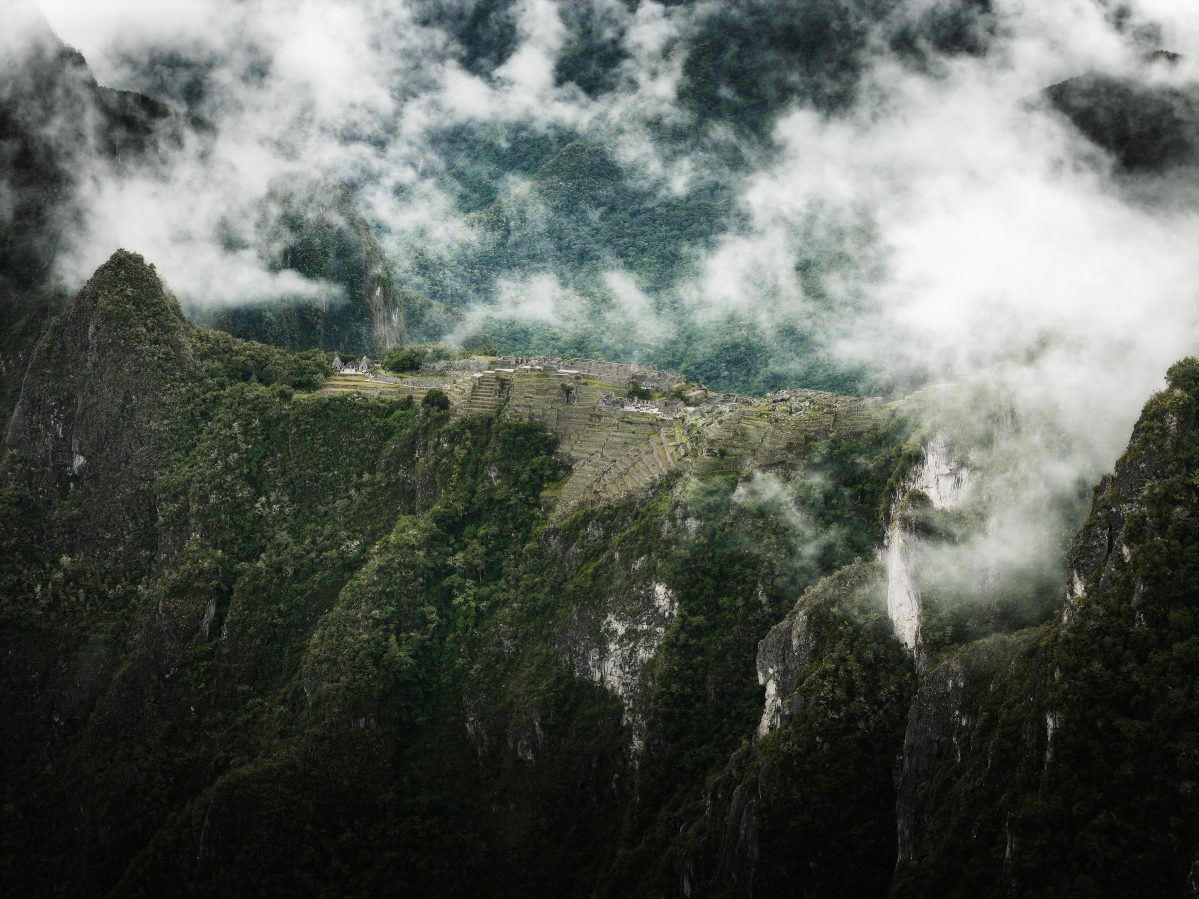Distant view of Machu Picchu terraces through clouds and forest