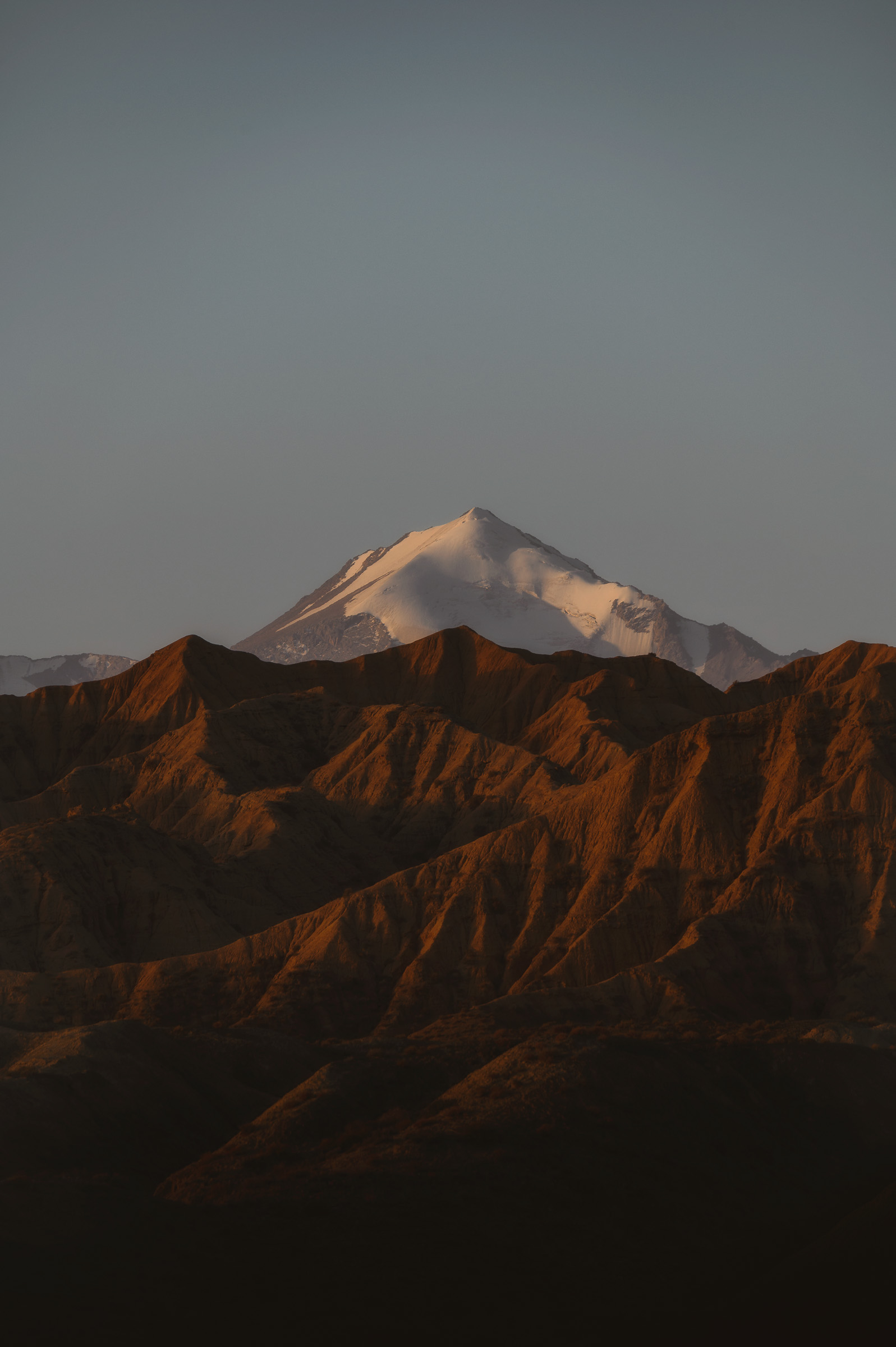 Snow-covered mountain above red ridges in evening light